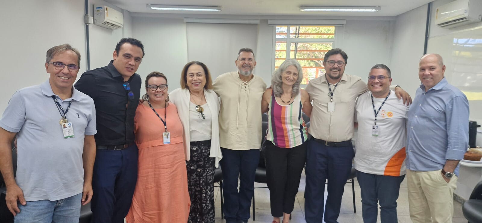 #ParaTodosVerem: Fotografia em plano médio, mais aproximada do mesmo grupo de nove pessoas da imagem anterior, posando juntas no mesmo ambiente de sala de reuniões. Eles aparecem alinhados e sorridentes, do peito/quadril para cima. A disposição das pessoas é exatamente a mesma: o homem de polo azul-clara na ponta esquerda, seguido pelo homem de camisa preta, a mulher de vestido coral, a mulher com casaco branco, o homem de bata bege centralizado, a mulher de blusa listrada colorida, o homem de camisa bege, o homem de camiseta branca e, na ponta direita, o homem de camisa social azul e calça bege.