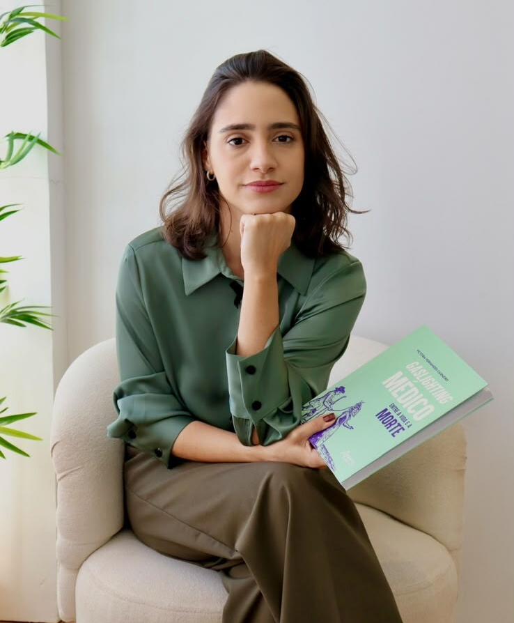 egressa_semifinalista#ParaTodosVerem: Fotografia de uma mulher jovem, de pele clara e cabelos castanhos curtos, sentada em uma poltrona arredondada de tecido off-white, em um ambiente interno bem iluminado. Ela está sentada de pernas cruzadas e olha para a câmera com uma expressão serena.