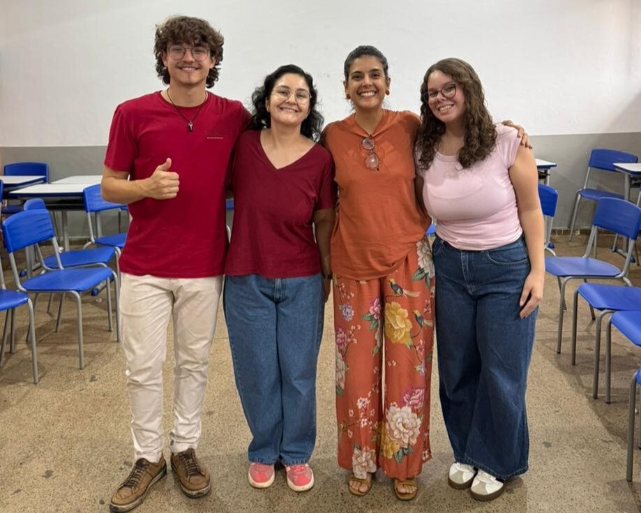 #ParaTodosVerem:Fotografia em plano inteiro de quatro pessoas em pé lado a lado, sorrindo, em uma sala de aula com cadeiras azuis ao fundo. Da esquerda para a direita: um homem de camiseta vermelha, calça clara e óculos, fazendo sinal de "joia" com a mão direita; uma mulher de camiseta vinho, calça jeans e óculos; uma mulher de blusa alaranjada e calça florida em tons de laranja e rosa; e uma mulher de camiseta rosa clara, calça jeans e óculos.