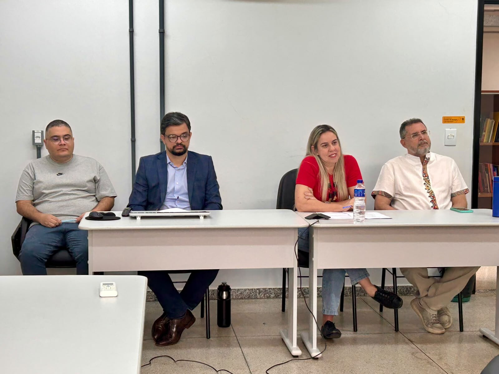 visita_intitucional_ppgdh#ParaTodosVerem:Quatro pessoas sentadas lado a lado atrás de uma mesa branca numa sala de reuniões. Da esquerda para a direita: homem de t-shirt cinzenta e óculos; homem de fato azul-marinho e óculos; mulher de t-shirt vermelha a falar; e homem de bata branca com bordados étnicos e óculos, a escutar atentamente.