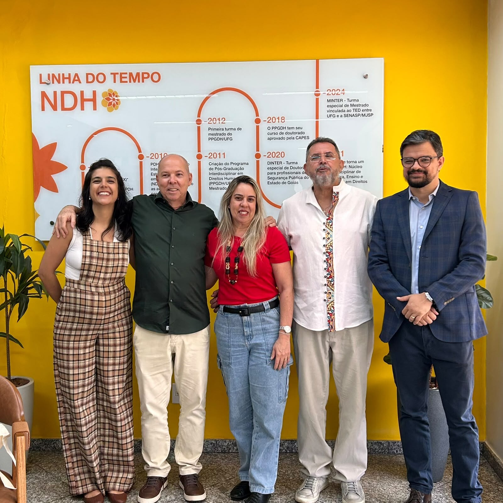 visita_intitucional_ppgdh#ParaTodosVerem:Cinco pessoas sorridentes alinhadas lado a lado em frente a um painel branco de linha do tempo numa parede amarela. Da esquerda para a direita: mulher em macacão xadrez, homem de camisa verde-escura, mulher de t-shirt vermelha e calça jeans, homem de bata branca com detalhes bordados e homem de fato azul-marinho.