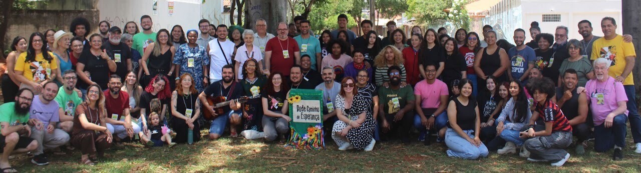 #ParaTodosVerem:Fotografia panorâmica em plano aberto de um numeroso grupo de pessoas reunidas ao ar livre, num espaço com relva e árvores ao fundo.O grupo está disposto em várias filas: algumas pessoas estão agachadas e sentadas na relva em primeiro plano, enquanto a maioria permanece em pé atrás. Vários participantes usam crachás ao pescoço e roupas casuais e coloridas. No centro, uma mulher de vestido preto a bolinhas brancas está ajoelhada ao lado de um estandarte verde decorado com fitas coloridas e girassóis, onde se lê "Rede Caminho de Esperança". À esquerda do estandarte, um homem de t-shirt azul toca violão. Todos sorriem e olham na direção da câmara num ambiente de confraternização.