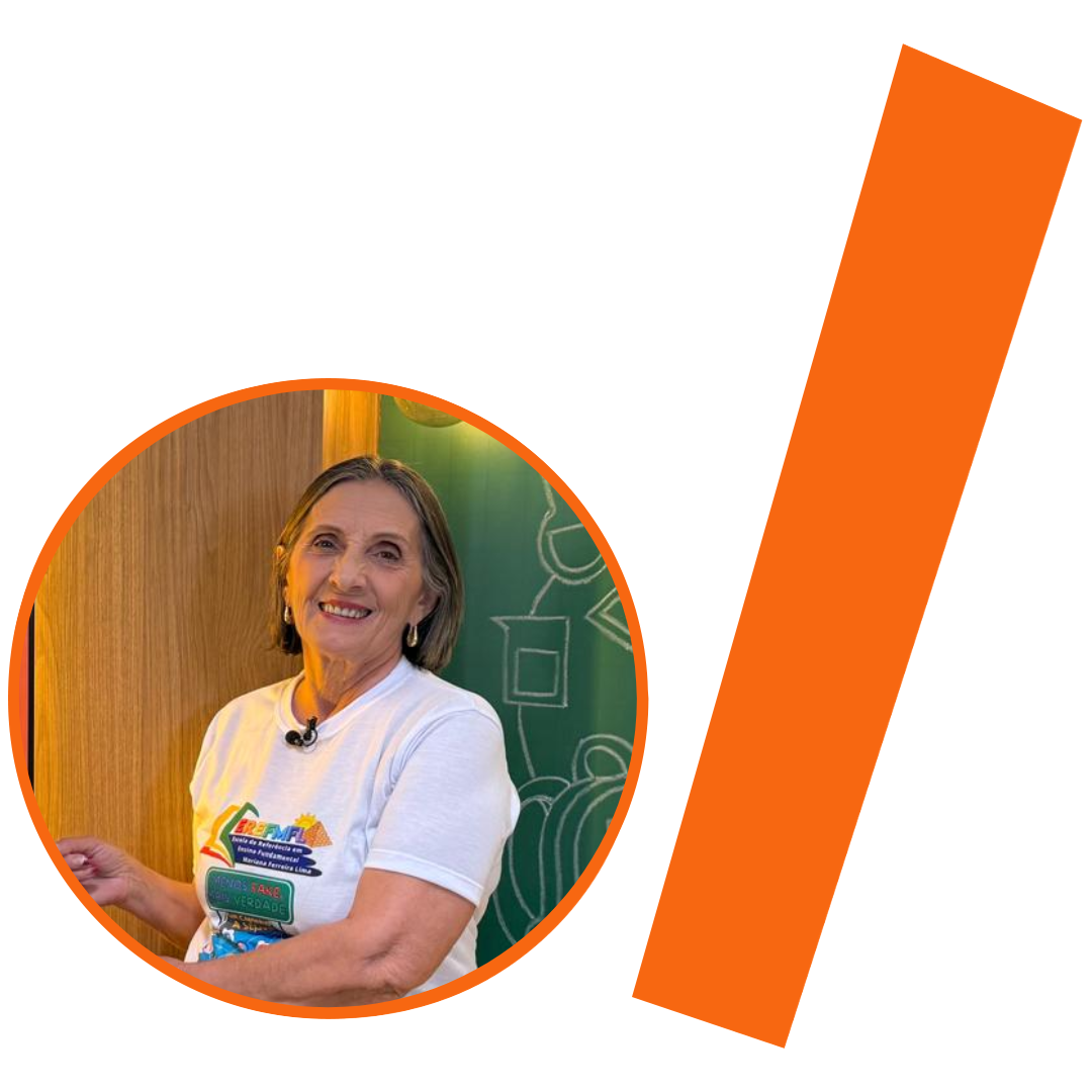 #ParaTodosVerem: Fotografia colorida de uma mulher branca idosa, com cabelos grisalhos curtos. Ela sorri para a câmera e veste uma camiseta branca de gola redonda com logotipos coloridos, incluindo um texto escrito "MENOS FAKE, MAIS VERDADE". A imagem está inserida em um recorte circular com borda laranja. Ao fundo da foto, vê-se um quadro-negro verde com desenhos abstratos em giz branco, além de parte de uma estrutura de madeira desfocada. À direita do círculo, há uma barra grossa e inclinada também na cor laranja. O conjunto visual está sobre um fundo branco liso, mantendo a identidade visual padrão do projeto.