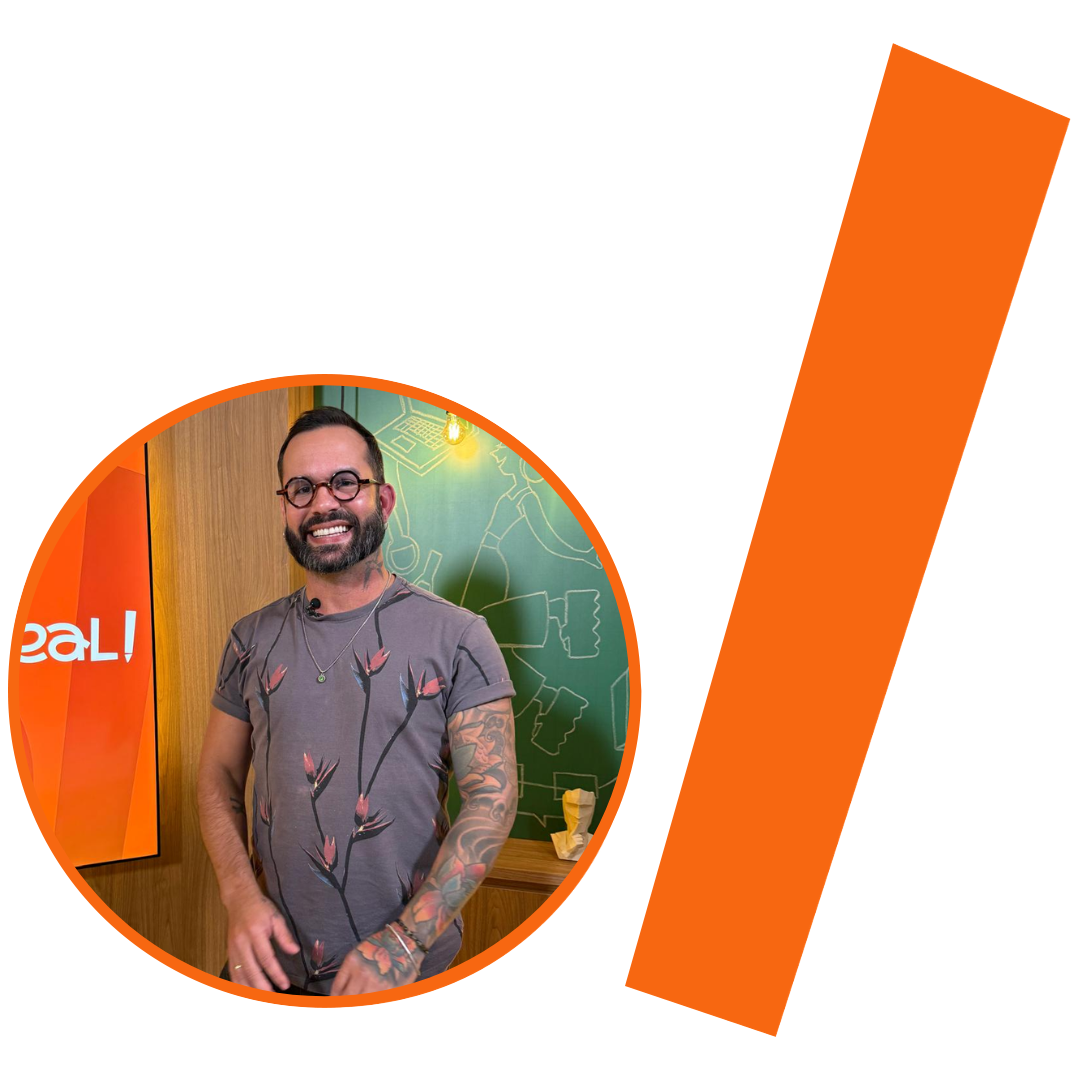 #ParaTodosVerem: Fotografia colorida em plano médio de um homem branco, com cabelos curtos e lisos, castanhos e barba cheia. Ele sorri para a câmera e usa óculos de grau redondos com armação de metal preta e colar de miçangas prateadas. Veste uma camiseta cinza-claro com estampa floral rosa e preta e uma calça de sarja preta. Ele tem tatuagens coloridas em ambos os braços. A imagem está inserida em um recorte circular com borda laranja. Ao fundo da foto, vê-se um quadro-negro verde com desenhos abstratos em giz branco, além de parte de uma estrutura de madeira desfocada e uma lâmpada de filamento pendurada e acesa. À direita do círculo, há uma barra grossa e inclinada também na cor laranja. 