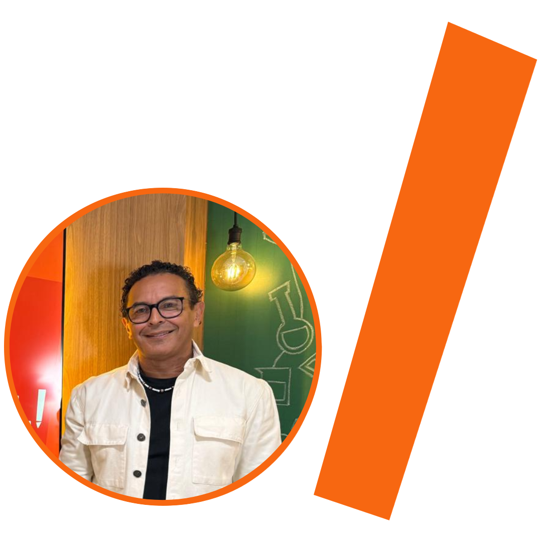 #ParaTodosVerem: Fotografia colorida em plano médio de um homem pardo, de cabelos pretos curtos e encaracolados, sorrindo. Ele usa óculos de grau com armação preta, um colar de contas pretas e brancas, e veste uma jaqueta de sarja branca sobre uma camiseta preta. Ao fundo, à esquerda, uma superfície de madeira e, à direita, uma parede verde com desenhos em giz e uma lâmpada de filamento acesa. A imagem está recortada em um formato circular com borda laranja. À direita do círculo, há uma barra grossa e inclinada também na cor laranja. O conjunto visual segue o padrão da identidade visual do projeto.