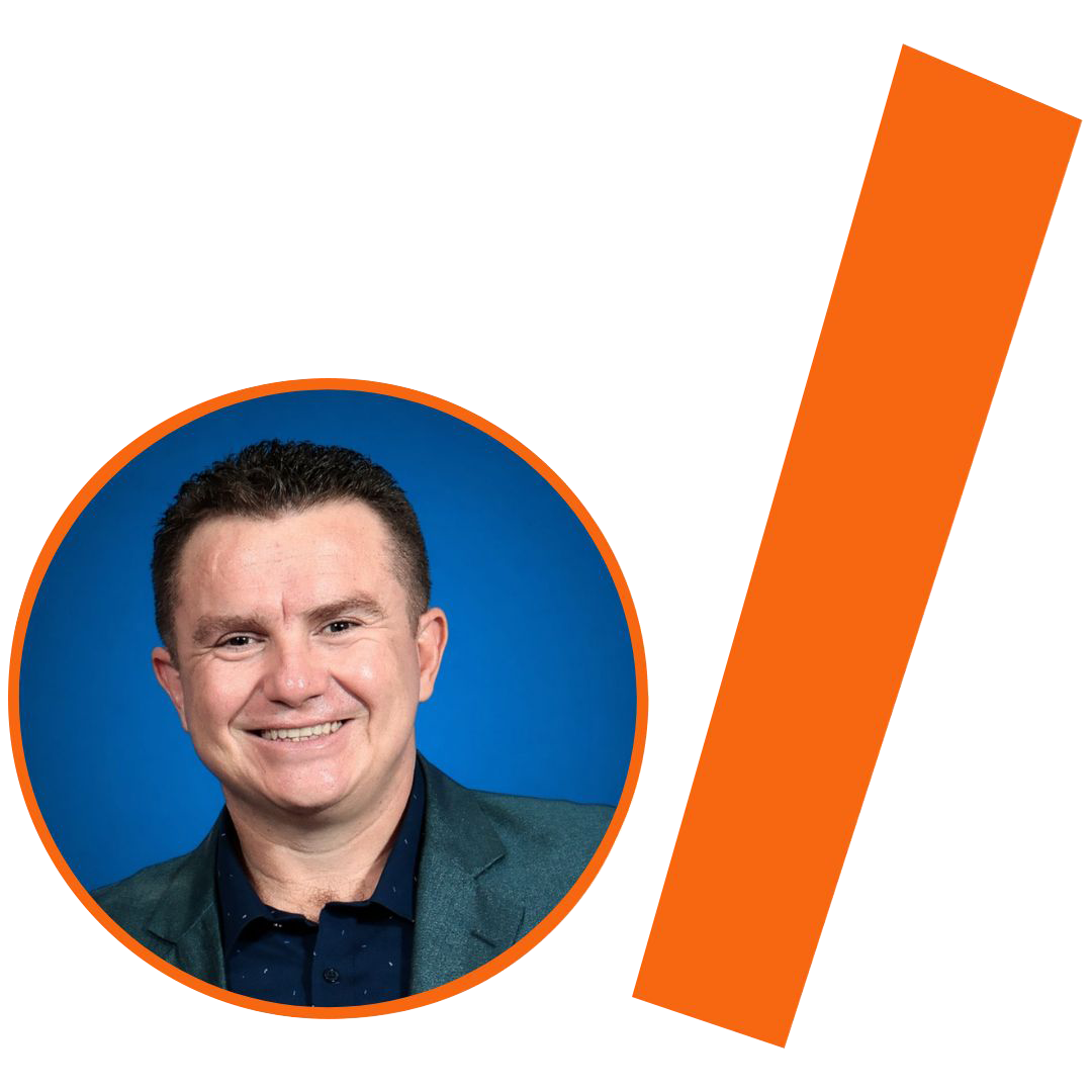 #ParaTodosVerem: Fotografia colorida em plano médio de um homem branco de meia-idade, com cabelos castanhos curtos e um sorriso largo. Ele veste um paletó cinza-esverdeado sobre uma camisa social azul-marinho com pequenos detalhes discretos. O fundo da foto é de um azul vibrante e uniforme. A imagem está recortada em um círculo com borda laranja. À direita do círculo, há uma barra grossa e inclinada também na cor laranja. O conjunto visual está mantendo o padrão da identidade visual do projeto.