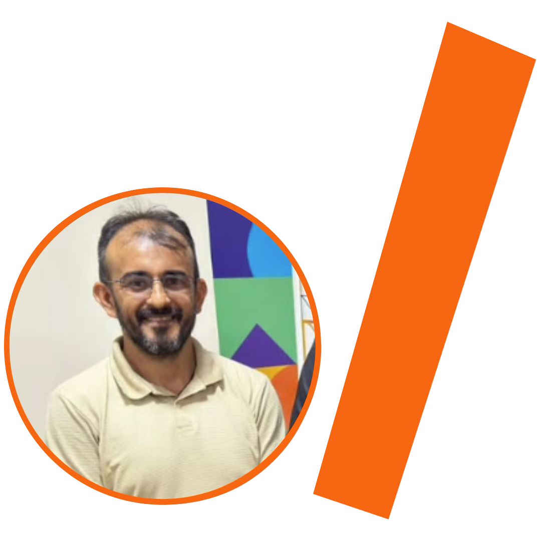 #ParaTodosVerem: Fotografia colorida em plano médio de um homem branco de meia-idade, com cabelos castanhos curtos e ralos e barba rala. Ele sorri para a câmera e usa óculos de grau com armação de metal fina. Veste uma camisa polo de cor bege claro. A imagem está inserida em um recorte circular com borda laranja. Ao fundo, uma parede branca e parte de um banner colorido com formas geométricas nas cores azul, verde e roxo. À direita do círculo, há uma barra grossa e inclinada também na cor laranja. O conjunto visual segue a identidade visual padrão do projeto.