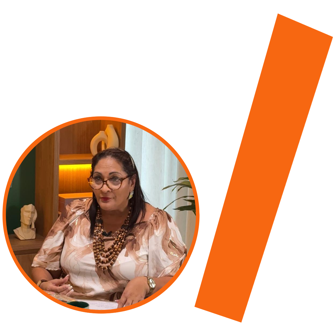 #ParaTodosVerem: Fotografia colorida em plano médio de uma parda branca de meia-idade, com cabelos pretos e lisos. Ela usa óculos de grau com armação grossa e escura, batom vermelho e vários colares de contas de madeira de diferentes comprimentos. Veste uma blusa estampada em tons de bege, marrom e branco. A imagem está inserida em um recorte circular com borda laranja. Ao fundo, um ambiente de escritório com estantes de madeira, objetos decorativos e uma planta. À direita do círculo, há uma barra grossa e inclinada na cor laranja. O conjunto visual está mantendo a identidade visual padrão do projeto.