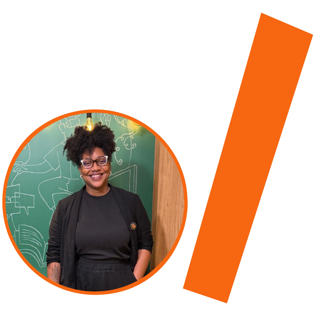 #ParaTodosVerem: Fotografia colorida em plano médio de uma mulher negra sorrindo, com cabelos pretos volumosos presos para cima. Ela usa óculos de grau com armação clara, um piercing no septo e brincos pequenos. Veste uma blusa preta e um cardigã da mesma cor, com um pequeno bottom circular na lapela. No antebraço direito, possui uma tatuagem. Ela está posicionada em um recorte circular com borda laranja. Ao fundo, uma parede verde com desenhos em giz branco e uma lâmpada de filamento acesa. À direita do círculo, há uma barra grossa e inclinada na cor laranja. A composição segue a identidade visual do projeto.