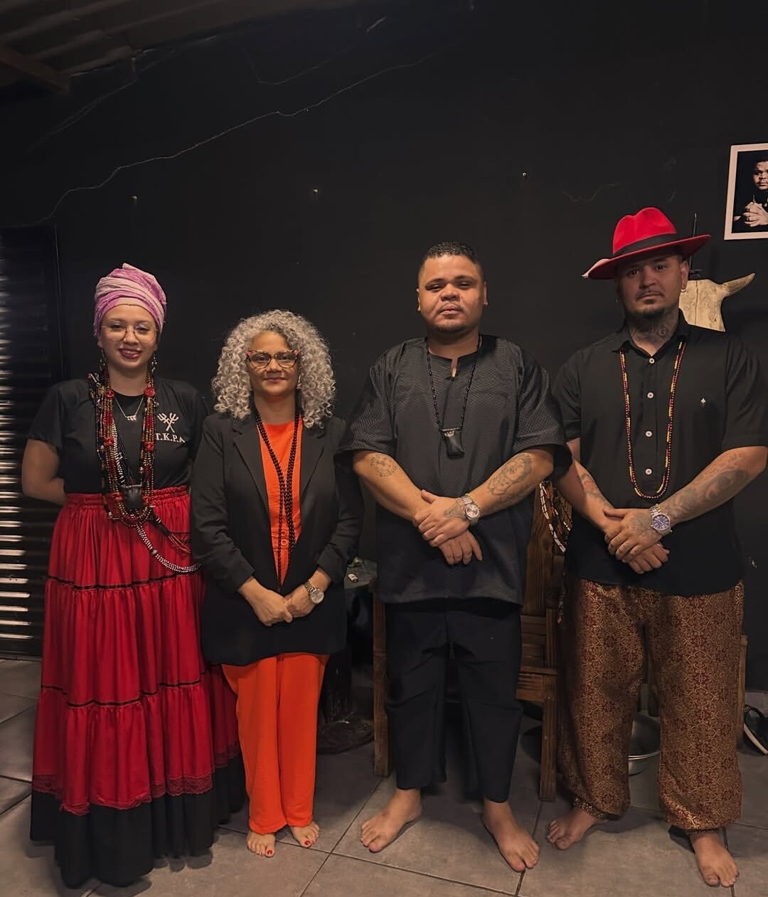 #ParaTodosVerem:Fotografia em plano inteiro de quatro pessoas descalças posando lado a lado em um ambiente de fundo escuro. Da esquerda para a direita: uma mulher com turbante lilás, saia vermelha e colares; uma mulher de cabelos grisalhos e blazer preto sobre roupa laranja; um homem de túnica preta; e um homem de chapéu vermelho, camisa preta e calça estampada.