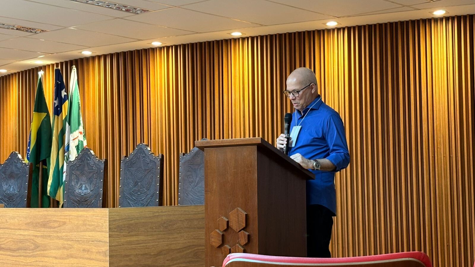 #ParaTodosVerem Foto de João da Cruz Gonçalves Neto, Professor da FD/UFG. Ele está de perfil, em pé atrás de um púlpito de madeira com a logomarca da UFG entalhada. Veste camisa azul royal e segura um microfone. Ao fundo, uma parede de madeira ripada e três bandeiras (Brasil, Goiás e UFG) em mastros de madeira esculpida.