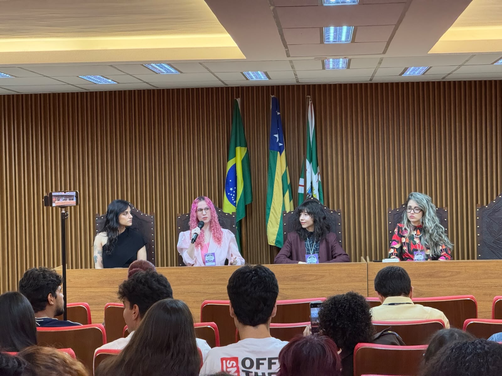 #ParaTodosVerem Foto do palco. Quatro juradas estão sentadas à mesa de madeira, de frente para a plateia (que aparece em primeiro plano, de costas). Atrás delas, as bandeiras do Brasil, Goiás e UFG. Da esquerda para a direita: uma jurada de preto; outra com cabelo rosa, falando ao microfone; ao seu lado outra jurada de blazer roxo; e a jurada de blusa floral colorida. Um smartphone em um tripé no canto esquerdo filma a cena.