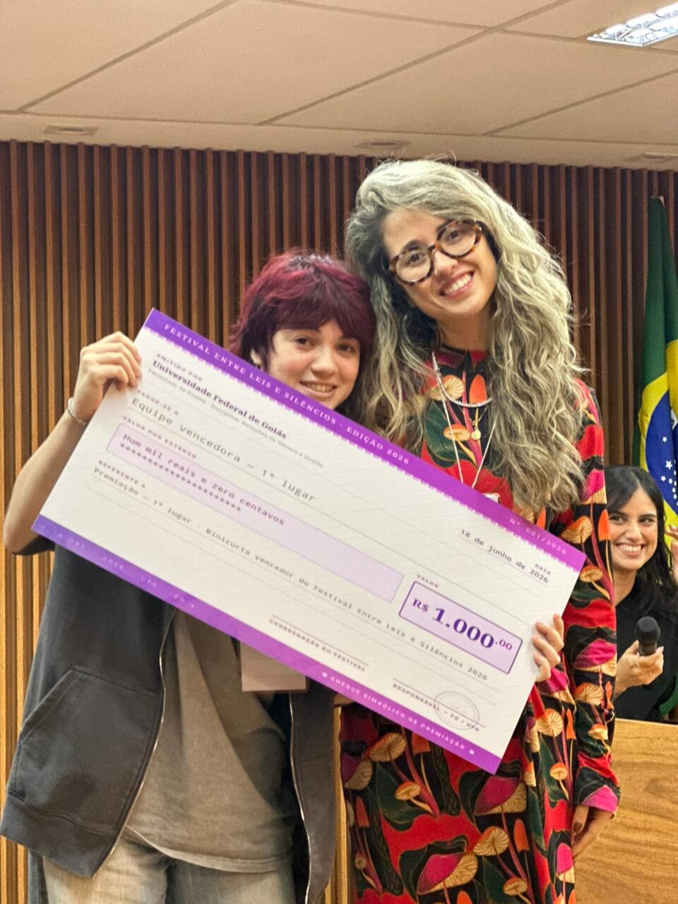 #ParaTodosVerem Foto da entrega do 1º Lugar. Duas pessoas sorriem segurando um grande cheque simbólico roxo e branco no valor de "R$ 1.000,00". À esquerda, a integrante da equipe vencedora, com cabelo ruivo curto e jaqueta escura. À direita, a jurada de blusa floral colorida e cabelos grisalhos cacheados. O cheque diz: "Festival Entre Leis e Silêncios - Edição 2026", "Equipe vencedora - 1º lugar". Ao fundo, a jurada sorri.