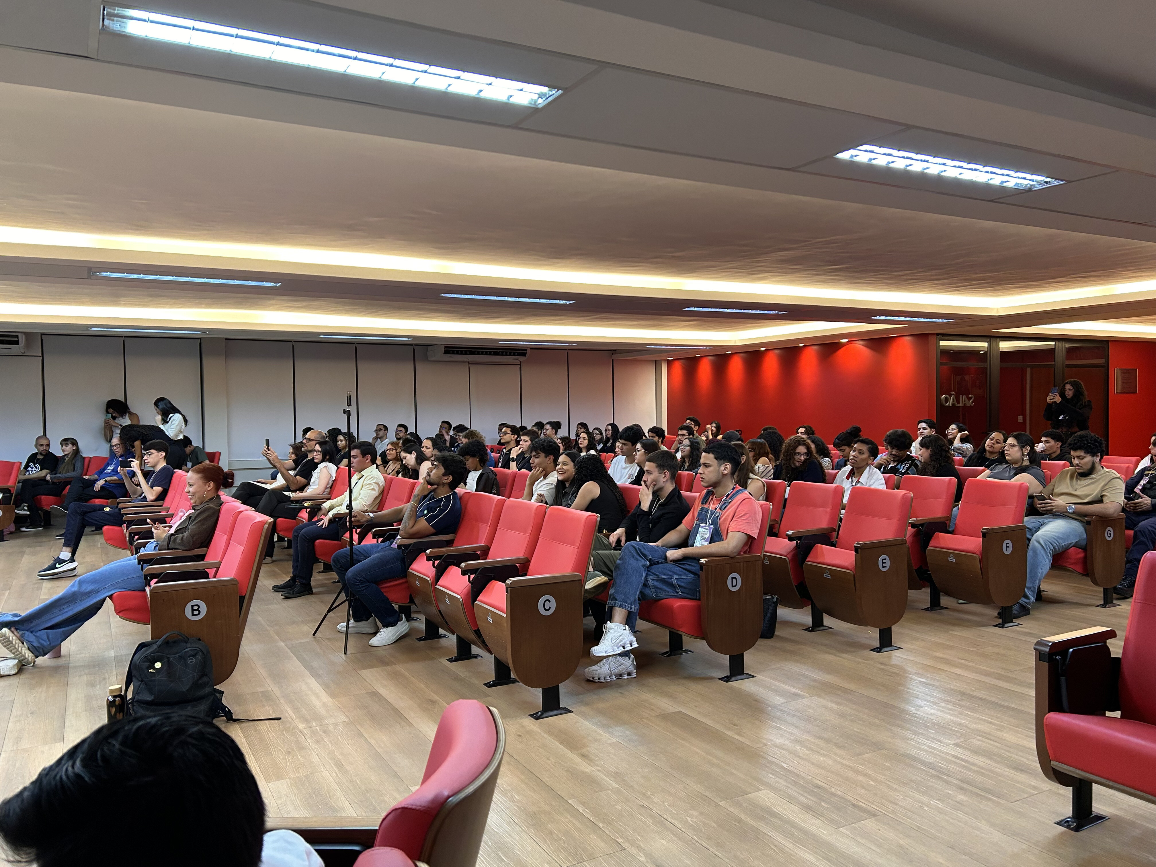 #ParaTodosVerem Foto panorâmica da plateia lotada no Salão Nobre. Dezenas de pessoas estão sentadas nas poltronas vermelhas, de costas ou perfil para a câmera, olhando para o palco (fora de quadro). Ao fundo, a parede vermelha e o letreiro iluminado "Salão Nobre FD - Faculdade de Direito".