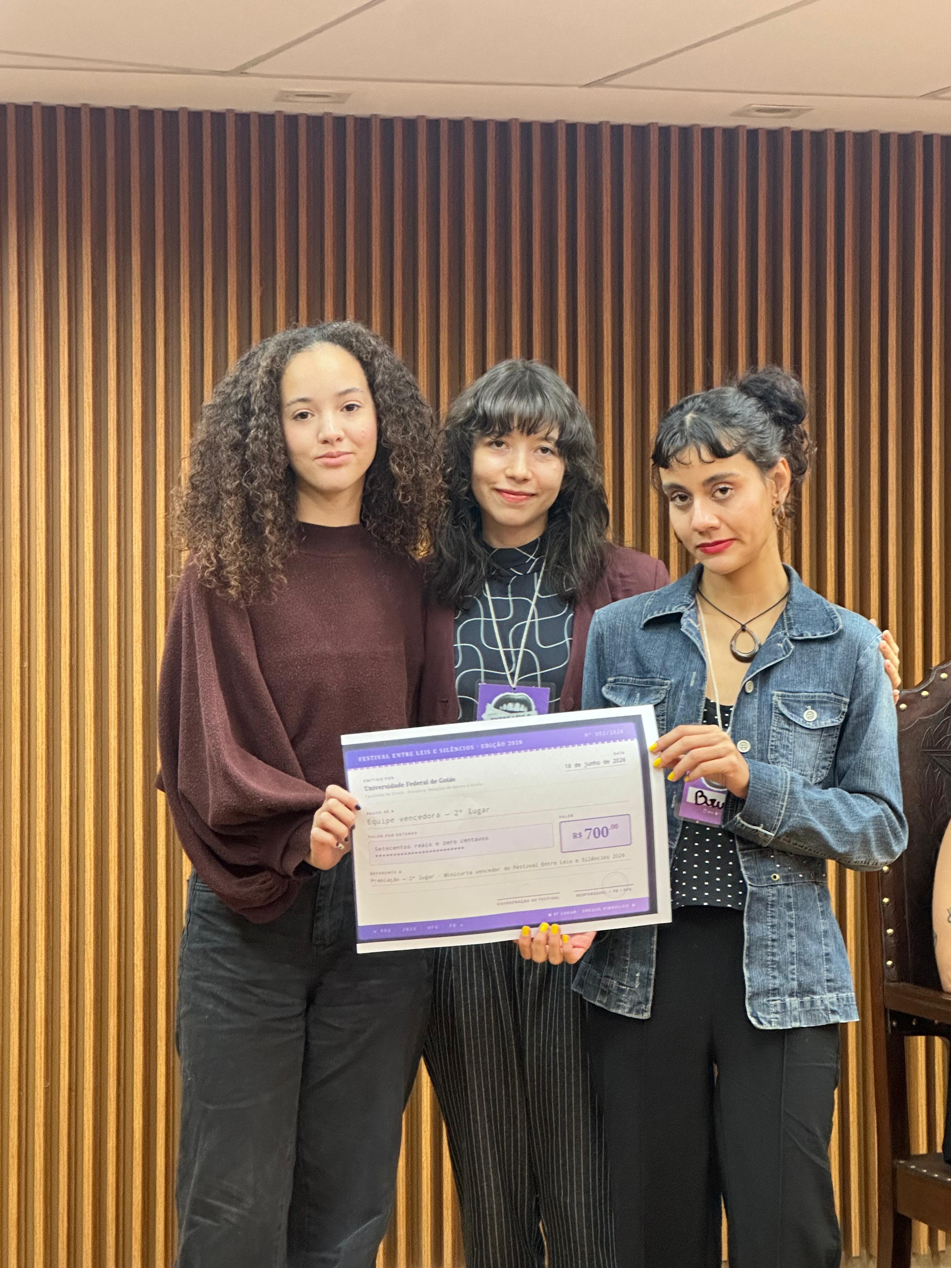 #ParaTodosVerem Foto da entrega do 2º Lugar. Três integrantes da equipe vencedora em pé, segurando o cheque simbólico de "R$ 700,00". Da esquerda para a direita: uma jovem de blusa roxa manga longa; uma jovem de blusa preta e azul com uma credencial pendurada; e uma jovem de jaqueta jeans. O cheque exibe as informações do festival e a colocação. Fundo de madeira ripada.