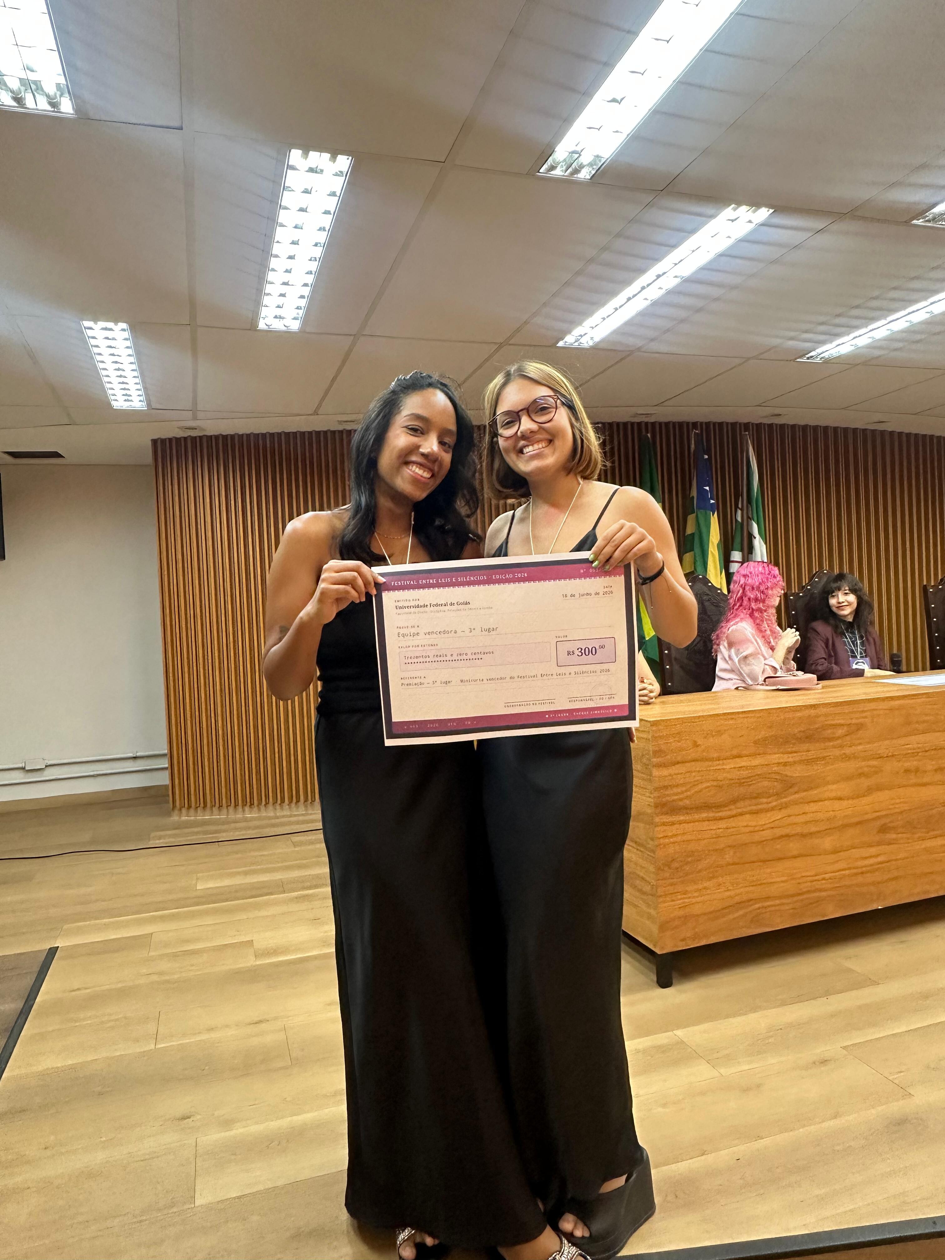 #ParaTodosVerem Foto da entrega do 3º Lugar. Duas integrantes da equipe vencedora, em pé, sorrindo e segurando o cheque simbólico de "R$ 300,00". Ambas vestem vestidos pretos longos de alça. O fundo é o palco com a mesa de madeira e as bandeiras.