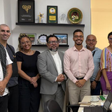 Banner_Notícia_CPDR - #ParaTodosVerem: Fotografia colorida de um grupo de oito pessoas em pé, sorrindo, em pose formal dentro de um escritório bem iluminado. Ao fundo, há duas prateleiras pretas fixadas na parede com diversos prêmios e troféus. À direita, um banner corporativo de roll-up verde e branco exibe o logotipo "DPE-GO" e o texto "Defensoria Pública do Estado de Goiás". As pessoas, que incluem homens e mulheres, vestem trajes variados, do casual ao profissional.
