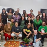 banner_noticia_cafe_com_direitos_humanos

#ParaTodosVerem Uma fotografia em grupo, colorida, de cerca de 25 pessoas de diversas idades, etnias e origens. Elas estão reunidas em quatro fileiras, algumas agachadas na frente, outras de pé atrás, sorrindo e olhando para a câmera. Estão num espaço interior amplo, que parece ser uma sala de aula ou um auditório, com paredes brancas, portas cinzentas e o que parecem ser mesas de formica visíveis nas bordas inferiores da imagem. No chão, à frente do grupo, há dois panos coloridos e estampados sobre um tapete menor e circular, com uma pequena planta em vaso no centro. As pessoas vestem uma variedade de roupas casuais e coloridas, incluindo t-shirts, blusas, vestidos e calças. Alguns têm óculos e chapéus. A iluminação é natural e difusa. A atmosfera geral é de calor e camaradagem.