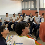 #ParaTodosVerem:

Fotografia de estudantes em uma sala de aula dispostos em círculo. Em primeiro plano e de costas, uma mulher de blusa alaranjada gesticula enquanto fala com os jovens. Ao redor, os alunos vestem uniforme escolar de camiseta branca com detalhes amarelos e azuis, sentados em cadeiras e sobre um tapete colorido no chão, todos atentos à conversa.