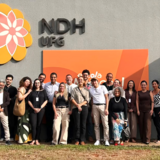 banner_notícia_visita_institucional_ppgdh
#ParaTodosVerem:

Fotografia em plano médio aproximado de um grupo de cerca de vinte pessoas alinhadas lado a lado e sorridentes, posando em frente ao edifício do NDH UFG.

Ao fundo, na parede cinza do prédio, destaca-se em grandes proporções a marca estilizada do NDH em formato de flor nos tons amarelo e rosa, ao lado do texto em relevo "NDH UFG" e de um painel laranja onde se lê parcialmente a palavra "escola". O grupo veste roupas casuais e formais em tons variados, como preto, bege, branco e estampado.