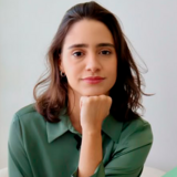 banner_notícia_semi-finalista
#ParaTodosVerem:

Retrato em primeiro plano de uma jovem de pele clara e cabelos castanhos curtos. Ela está ligeiramente inclinada para a frente, com o queixo apoiado na mão e um semblante sereno, olhando para a câmera. Veste uma camisa de cetim verde-oliva. Ao fundo, uma parede branca iluminada e, no canto esquerdo, folhas de uma planta verde. No canto inferior direito, aparece a ponta de um livro verde-claro.