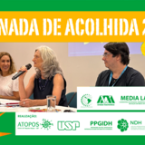 #ParaTodosVerem: Banner retangular com moldura verde. No topo, sobre uma larga faixa amarela, está escrito em letras brancas: "JORNADA DE ACOLHIDA 2026". No centro, há uma fotografia de três pessoas sentadas à mesa de um evento: à esquerda, uma mulher loira sorri; no centro, uma mulher de cabelos grisalhos fala ao microfone; e à direita, um homem de óculos observa. Sobreposta à direita da foto, há uma colagem com uma mão em preto e branco segurando a ilustração de uma lâmpada amarela acesa. No rodapé, sobre uma faixa branca, alinham-se em verde as logomarcas das instituições organizadoras e parceiras, como USP, UFG, Media Lab, entre outras.
