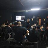 #ParaTodosVerem:

Fotografia em plano aberto de uma palestra em um galpão com paredes pretas e teto escuro, iluminado por lâmpadas no centro. Uma mulher de cabelos volumosos e grisalhos fala em pé, ao fundo, ao lado de uma tela de projeção ligada com o título "Mediar é ancestral".

A plateia está disposta em semicírculo em primeiro plano e vista de costas, sentada em bancos de madeira e cadeiras plásticas escuras. Nas paredes pretas do espaço, há crânios decorativos de bois pendurados.