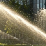 Imagem de um sistema de irrigação por aspersão instalado em área verde urbana, em frente a um edifício comercial. O equipamento é utilizado para manter jardins e gramados irrigados com eficiência, podendo operar com água de reúso em práticas sustentáveis de manejo hídrico.