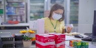 A foto mostra o interior de uma farmácia ou dispensário de medicamentos. Em primeiro plano, sobre o balcão, há várias caixas de remédios empilhadas, incluindo caixas vermelhas com a marca “ÉTIRA levetiracetam” e outras caixas coloridas à direita. Também há um pequeno vasinho com duas flores amarelas artificiais.

Ao fundo, um pouco desfocada, há uma mulher usando máscara facial e camiseta amarela. Ela está manipulando uma caixa de medicamento enquanto parece conferir informações em um papel. Atrás dela há prateleiras com diversos produtos e medicamentos, e à direita vê-se parte de um computador.

O ambiente é claro e organizado, com uma divisória transparente entre o balcão e a área de atendimento.
