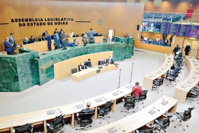 A imagem mostra o plenário da Assembleia Legislativa do Estado de Goiás durante uma sessão. Vários parlamentares e assessores circulam e conversam na área superior, enquanto alguns ocupam seus assentos na parte inferior. O espaço é amplo, com mesas curvas, cadeiras e painéis eletrônicos ao fundo.