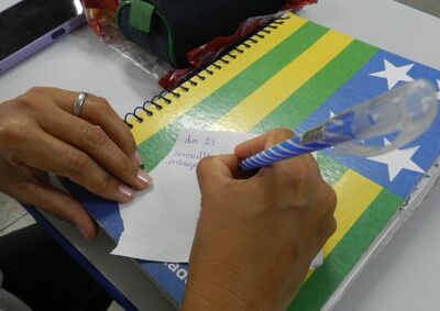 A foto mostra uma pessoa escrevendo em um pequeno pedaço de papel apoiado sobre um caderno espiralado. O caderno tem uma capa colorida com listras verdes, amarelas e azuis, além de estrelas brancas — remetendo à bandeira do estado de Goiás. A pessoa segura uma caneta azul com listras brancas e usa um anel prateado na mão esquerda. No papel, é possível ler parte de um lembrete escrito à mão: “dia 23 …”.