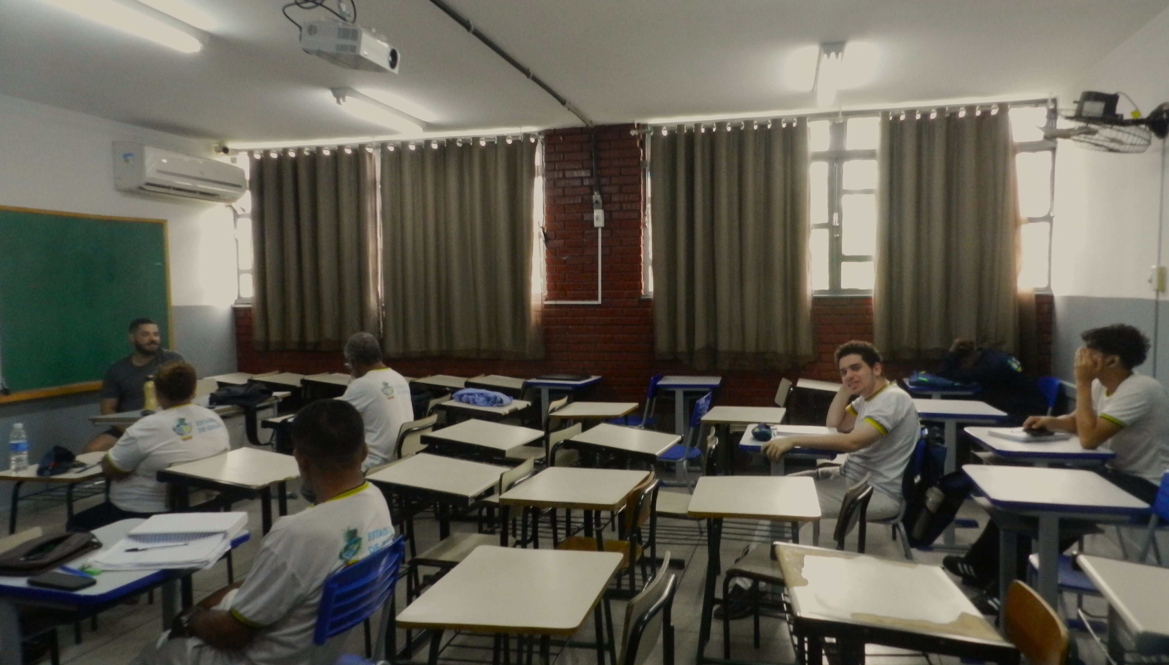 Sala de aula com várias carteiras distribuídas de forma organizada, algumas vazias e outras ocupadas por estudantes usando uniformes brancos. À esquerda, um homem sentado parece conduzir a atividade. As janelas ao fundo estão cobertas por cortinas bege e a sala é iluminada por lâmpadas fluorescentes. Um projetor está instalado no teto.
