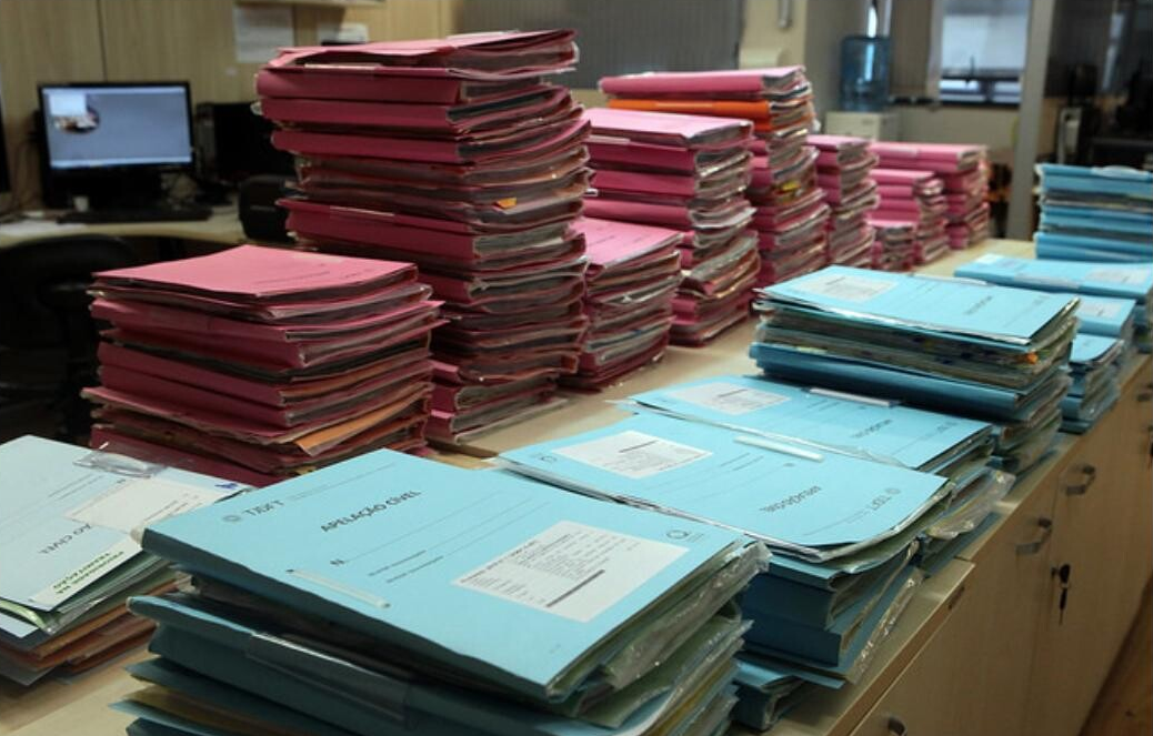 Fotografia de um ambiente de escritório (possivelmente um cartório ou setor judicial) mostrando uma grande quantidade de pilhas de processos e documentos físicos empilhados sobre uma mesa.Primeiro Plano: Dominado por pilhas de pastas processuais de cor azul-claro (ou ciano), algumas rotuladas como "APELAÇÃO CÍVEL". Estas pastas estão empilhadas em várias colunas baixas, ocupando a maior parte do centro e da direita da mesa.Segundo Plano (Fundo): Várias pilhas muito altas de processos ou volumes de documentos, majoritariamente envoltos em capas de cor rosa forte (ou magenta). Essas pilhas se estendem horizontalmente ao longo do centro da imagem, sugerindo um alto volume de trabalho ou acervo.Contexto: Ao fundo, é possível ver um monitor de computador em uma mesa de trabalho escura, sugerindo um contraste entre a documentação física (papel) e o ambiente de trabalho moderno.