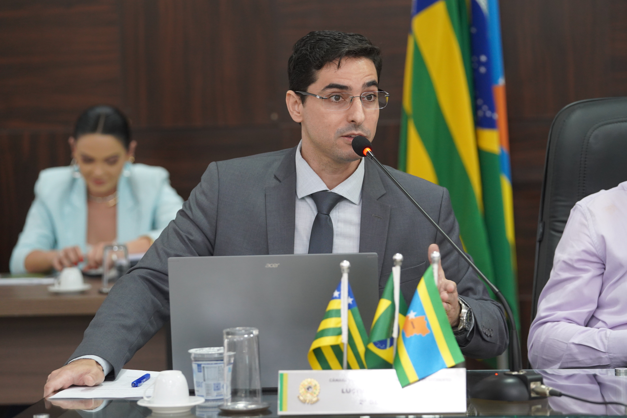 A imagem mostra um homem, vestido com terno e gravata, falando ao microfone durante uma sessão ou reunião oficial. Ele está sentado diante de um notebook, com bandeiras de Goiás e do Brasil ao lado, e parece participar de uma audiência ou comissão.