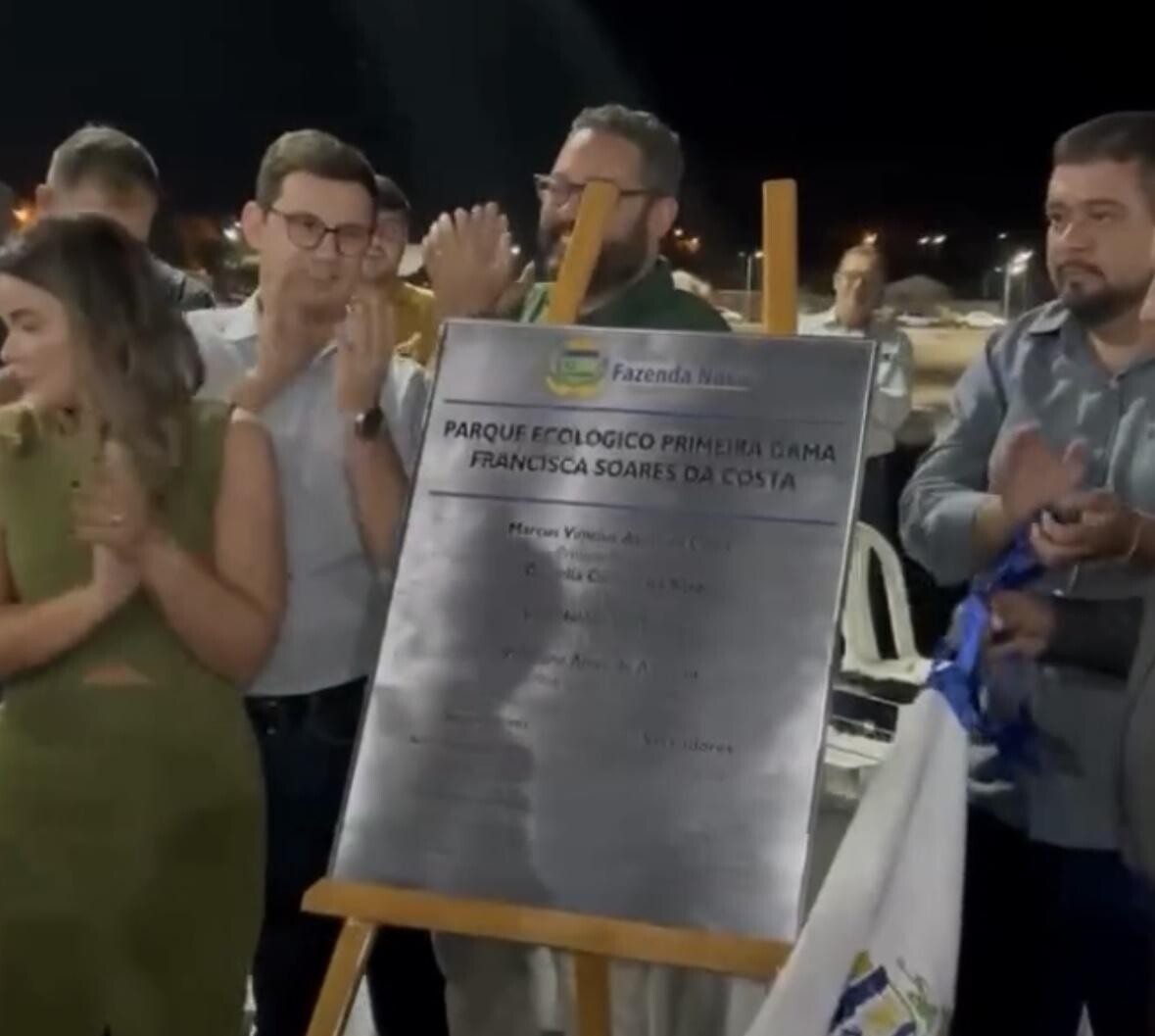 A imagem mostra um grupo de pessoas aplaudindo durante a inauguração de uma obra pública. No centro, há uma placa indicando a abertura do “Parque Ecológico Primeira Dama Francisca Soares da Costa”, em Fazenda Nova. A cena ocorre à noite, em ambiente externo.