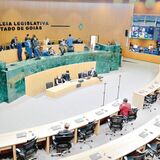 A imagem mostra o plenário da Assembleia Legislativa do Estado de Goiás durante uma sessão. Vários parlamentares e assessores circulam e conversam na área superior, enquanto alguns ocupam seus assentos na parte inferior. O espaço é amplo, com mesas curvas, cadeiras e painéis eletrônicos ao fundo.