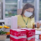 A foto mostra o interior de uma farmácia ou dispensário de medicamentos. Em primeiro plano, sobre o balcão, há várias caixas de remédios empilhadas, incluindo caixas vermelhas com a marca “ÉTIRA levetiracetam” e outras caixas coloridas à direita. Também há um pequeno vasinho com duas flores amarelas artificiais.

Ao fundo, um pouco desfocada, há uma mulher usando máscara facial e camiseta amarela. Ela está manipulando uma caixa de medicamento enquanto parece conferir informações em um papel. Atrás dela há prateleiras com diversos produtos e medicamentos, e à direita vê-se parte de um computador.

O ambiente é claro e organizado, com uma divisória transparente entre o balcão e a área de atendimento.