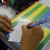 A foto mostra uma pessoa escrevendo em um pequeno pedaço de papel apoiado sobre um caderno espiralado. O caderno tem uma capa colorida com listras verdes, amarelas e azuis, além de estrelas brancas — remetendo à bandeira do estado de Goiás. A pessoa segura uma caneta azul com listras brancas e usa um anel prateado na mão esquerda. No papel, é possível ler parte de um lembrete escrito à mão: “dia 23 …”.