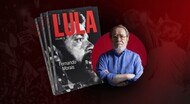 Arte de divulgação com fundo em tons de vermelho escuro. À esquerda, aparecem várias cópias empilhadas do livro Lula – Volume 2, de Fernando Morais. A capa destaca uma fotografia em preto e branco do rosto do presidente Luiz Inácio Lula da Silva em perfil, com o título “LULA” em letras grandes vermelhas.

À direita, há a imagem do escritor Fernando Morais, um homem idoso de cabelos grisalhos, barba curta e óculos. Ele veste uma camisa azul de mangas compridas e está com os braços cruzados, olhando para a câmera. Atrás dele, um círculo vermelho destaca sua figura.

O conjunto da imagem tem um visual sóbrio e institucional, remetendo ao lançamento da nova biografia de Lula escrita por Fernando Morais.