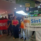 A imagem mostra um grupo de pessoas em uma manifestação dentro de um aeroporto ou rodoviária. No primeiro plano, há uma mulher de óculos escuros e blusa rosa ao lado de um homem de camiseta branca e laranja. Eles estão perto de uma grade com faixas do sindicato SINT-IFESgo. Acima deles, um cartaz diz: “Não é reforma, é o fim do serviço público”. Ao fundo, outras pessoas seguram uma grande faixa vermelha da CUT.