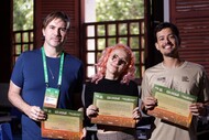 Fotografia de três pessoas em pé, posando em uma sala com janelas de madeira ao fundo. Elas sorriem e seguram certificados de participação em uma atividade formativa do FICA 2025. A pessoa à esquerda usa camiseta azul e crachá verde; ao centro, uma pessoa de cabelo rosa e óculos segura o certificado; à direita, uma pessoa com camiseta bege também exibe o certificado. A imagem registra um momento de conclusão e reconhecimento de participantes em uma ação educativa ou cultural.