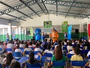 Fotografia de uma apresentação educativa em um espaço coberto, com várias crianças sentadas em cadeiras azuis, de costas para a câmera. À frente, uma pessoa fala ao microfone ao lado de dois personagens fantasiados, um azul e outro laranja. O ambiente tem banners da Saneago, cartazes sobre água e saneamento, caixas de som e estrutura metálica no teto.