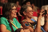 Fotografia de duas mulheres sentadas em uma plateia com poltronas vermelhas. A mulher à esquerda usa blusa verde e óculos, sorrindo enquanto assiste à atividade. A mulher à direita usa óculos, blusa estampada e segura um celular, aparentemente registrando a cena. Ao fundo, outras pessoas aparecem desfocadas na plateia.