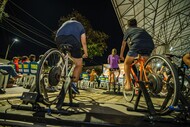 Fotografia noturna de pessoas pedalando em bicicletas ergométricas instaladas em uma área externa coberta. Em primeiro plano, aparecem duas bicicletas vistas por trás, com participantes pedalando. Ao fundo, há pessoas sentadas em cadeiras, iluminação urbana e uma estrutura coberta, sugerindo uma atividade coletiva ao ar livre.