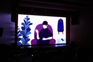Fotografia de uma sala de cinema escura durante uma exibição. Na tela grande, aparece uma cena de animação com uma pessoa sentada em uma poltrona roxa, uma planta à esquerda e uma mochila à direita. Na parte inferior da imagem, vê-se a silhueta do público sentado nas poltronas.