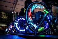 Fotografia em close de rodas de bicicletas ergométricas em movimento, iluminadas por luzes coloridas em tons de azul, verde, vermelho e roxo. As rodas aparecem enfileiradas em uma área escura, criando efeito visual luminoso durante a pedalada.