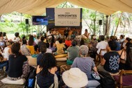 Registro de um painel ou roda de conversa realizado sob uma tenda, em ambiente externo e arborizado. Ao centro, há pessoas sentadas em cadeiras participando da discussão, com microfones e estrutura de som. O público acompanha a atividade sentado em cadeiras, almofadas e bancos, disposto em semicírculo. Ao fundo, aparece um painel com a identificação “Fórum Horizontes — Diálogos para novos caminhos” e uma tela lateral com a marca do FICA 2026.