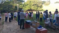 Grupo de crianças e adultos reunidos ao ar livre, em uma área com árvores, gramado e calçada de pedra. Algumas pessoas usam camisetas com identificação institucional, e há caixas e equipamentos apoiados sobre bancos próximos ao grupo. A cena mostra os participantes em roda, aparentemente acompanhando uma explicação ou atividade.