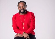 Retrato em estúdio de um homem sorrindo, sentado, usando suéter vermelho, calça escura e acessórios dourados, como colar com pingente de cruz, pulseira e anéis. O fundo é claro e neutro, com iluminação suave.