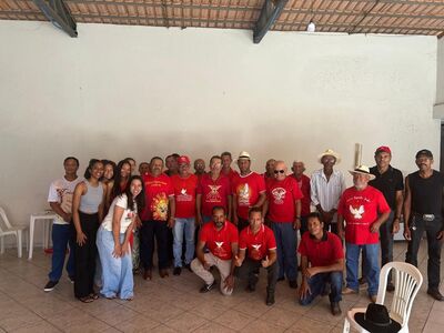 Grupo de pessoas posando em um salão coberto, diante de uma parede clara. Parte do grupo veste camisetas vermelhas com estampas brancas, enquanto outras pessoas usam roupas casuais. Algumas estão em pé e outras agachadas à frente. O espaço tem piso de cerâmica, cadeiras plásticas nas laterais e telhado aparente.